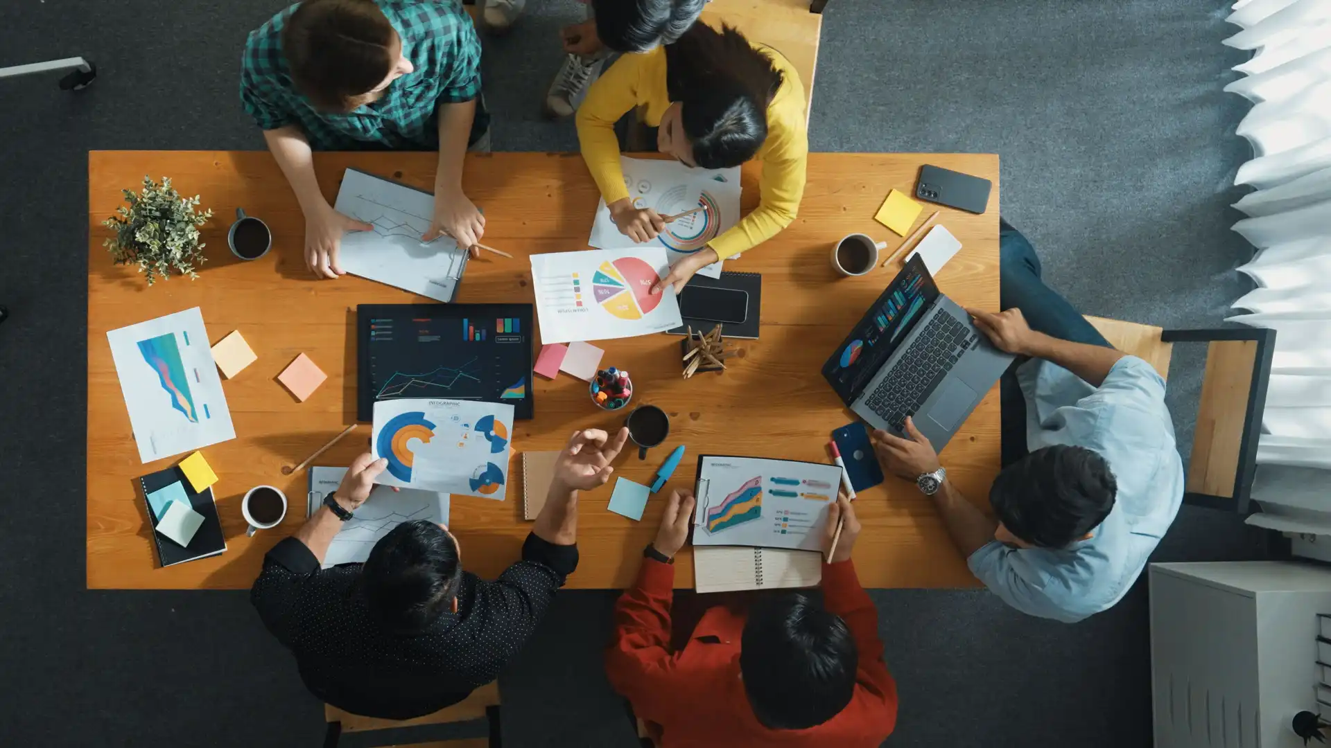 Foto de profissionais de marketing discutindo sobre planejamento estratégico, com base nos resultados apresentados em gráficos de diversos formatos espalhados em cima de uma ampla mesa, no escritório físico da agência de marketing em que trabalham.