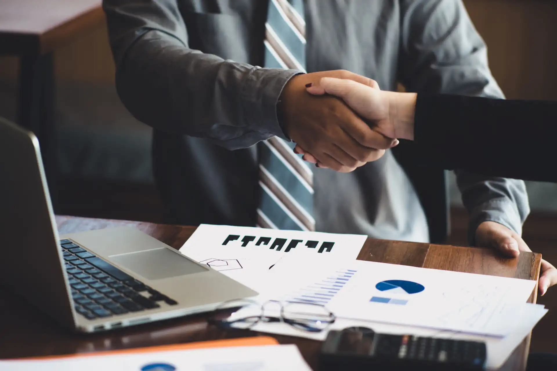 Foto de um aperto de mão entre duas pessoas de etnias e gêneros diferentes, em cima de uma mesa com diversos gráficos, papéis e calculadora espalhados, representando uma venda complexa em uma negociação B2B bem-sucedida.