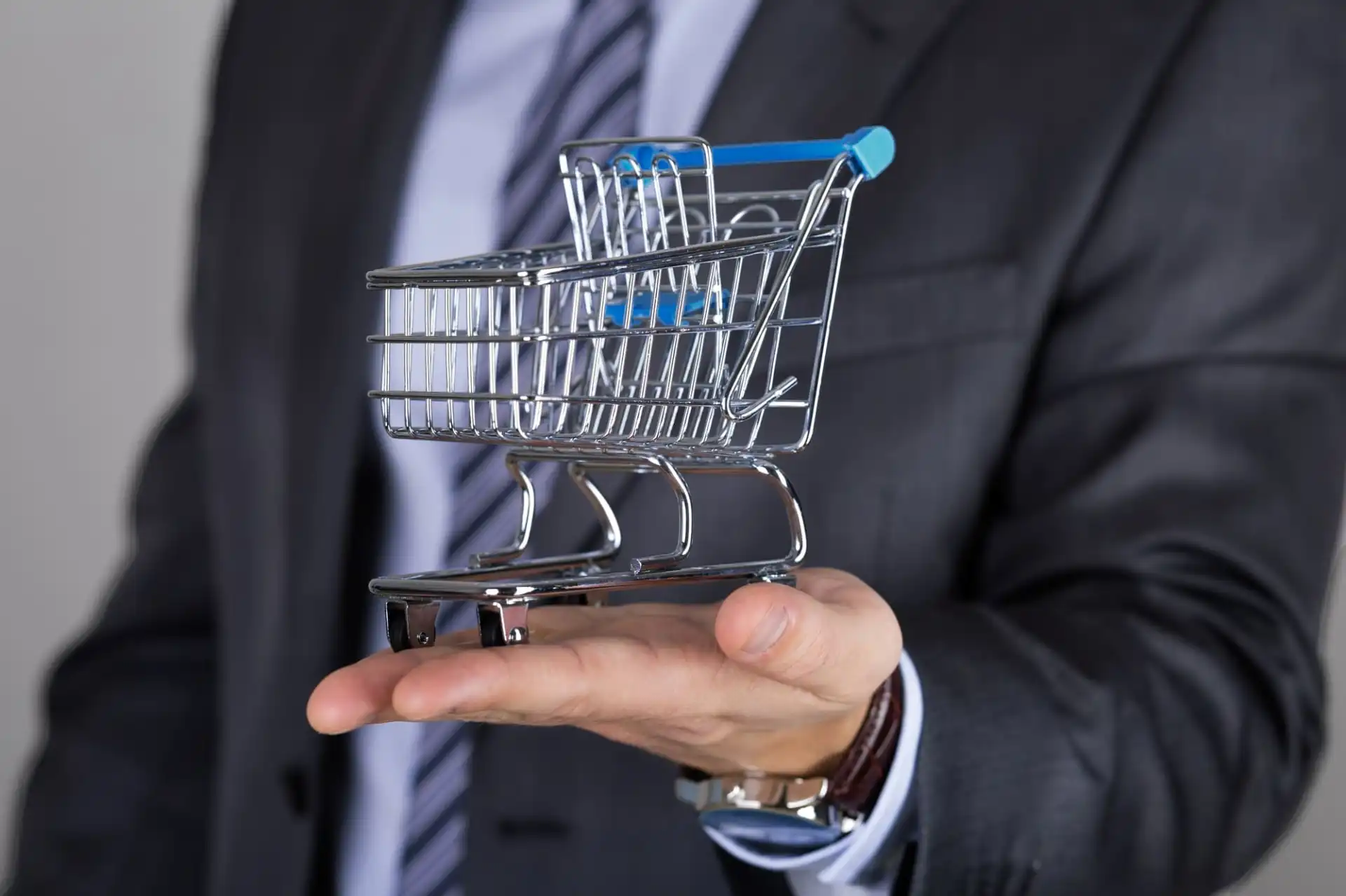 Foto do tronco de um homem de terno segurando a miniatura de um carrinho de compras na palma da mão esquerda, representando uma compra corporativa.