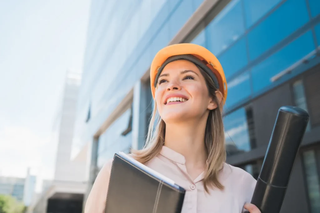 Foto de uma mulher engenheira de capacete de segurança sorrindo em frente a um prédio, representando atração de talentos via cases de autoridade.