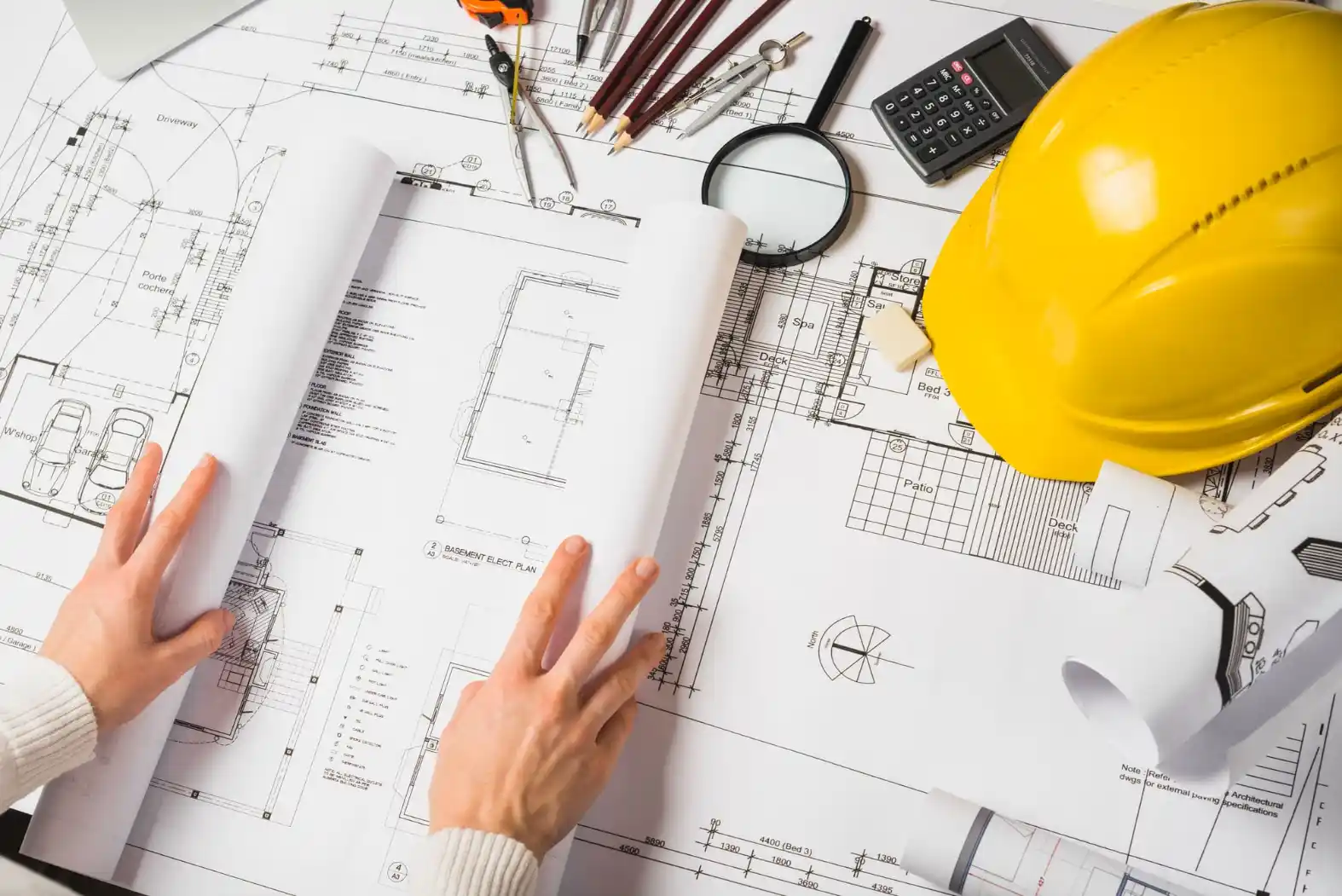 Foto de projetos de construção civil com plantas, ferramentas de desenho, uma calculadora, uma lanterna e um capacete de segurança amarelo.