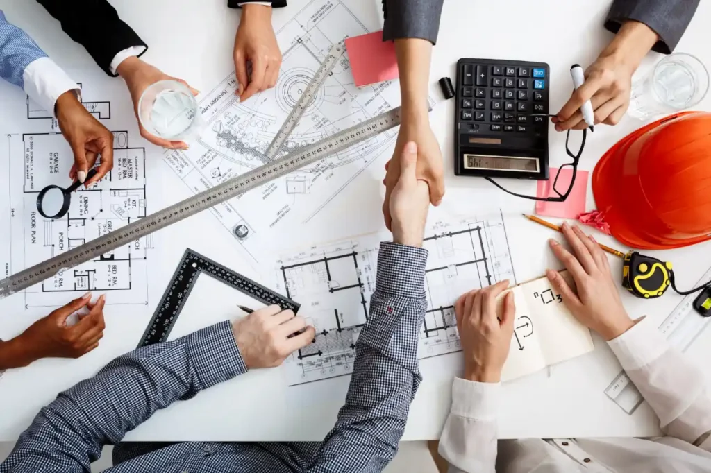 Vista superior de uma mesa de trabalho com profissionais de engenharia e construção civil analisando plantas arquitetônicas e documentos técnicos. Diversas mãos seguram réguas, calculadora, lupa e materiais de escritório enquanto outras duas mãos se apertam, sinalizando uma contração fechada.