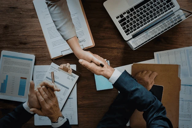 Foto de duas pessoas de gêneros diferentes apertando as mãos em uma mesa de trabalho com laptops, documentos, um contrato assinado e gráficos financeiros ao redor, simbolizando parceria e negócios potencializados com a estratégia de Marketing de Relacionamento B2B.