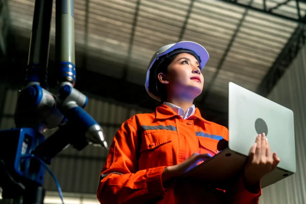 Foto, em perspectiva baixa, de uma jovem engenheira de capacete e uniforme laranja em ambiente industrial, usando tablet e observando um equipamento com atenção.