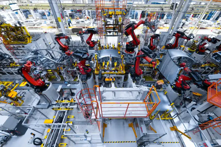 Foto da vista aérea de uma linha de produção industrial automatizada, com robôs articulados em vermelho e amarelo, braços mecânicos e máquinas em um grande galpão de fábrica.