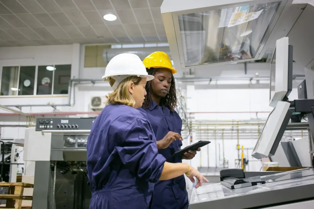 Foto de duas mulheres usando capacetes e uniformes de trabalho em uma planta industrial, analisando informações em um painel de controle ao lado de uma máquina grande.