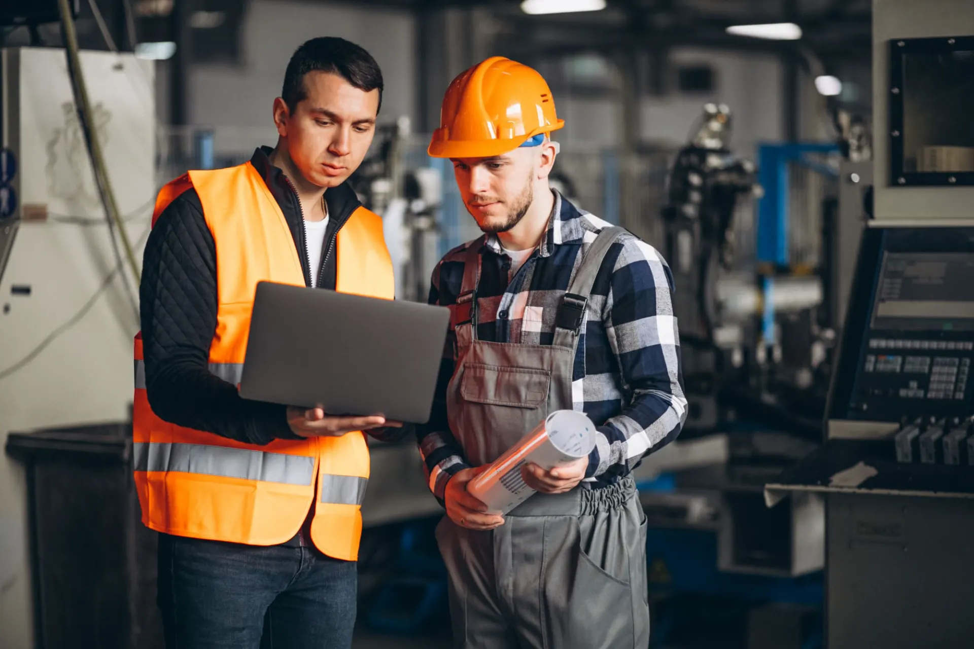 Foto de dois trabalhadores de indústria em ambiente de fábrica usam coletes e capacete de segurança, um deles segura um documento e o outro analisa um notebook.