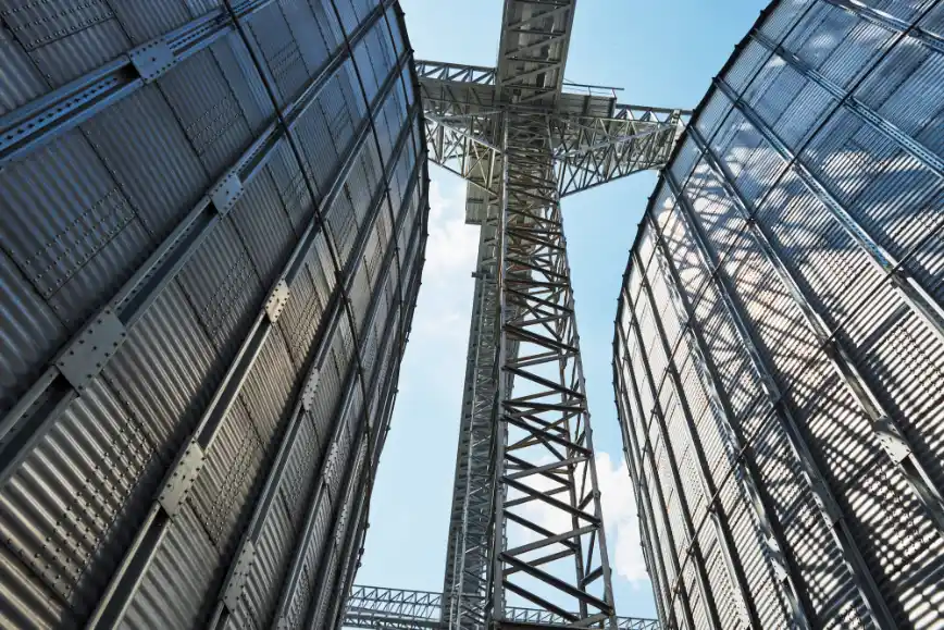 Foto da vista em ângulo baixo de silos metálicos com estruturas de aço, telhados corrugados e torre central de suporte, em ambiente industrial ao ar livre.
