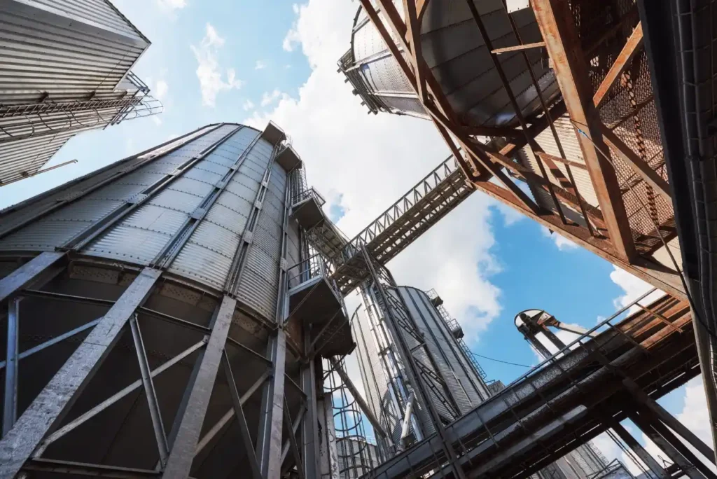 Foto da vista em ângulo baixo de um complexo industrial com silos metálicos, estrutura de aço e tubulações, com céu azul ao fundo.