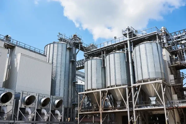 Foto da vista externa de uma planta industrial com silos de armazenamento e tubulações metálicas, conjunto de equipamentos e estruturas em uma fábrica.