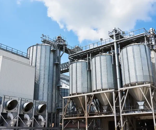 Foto da vista externa de uma planta industrial com silos de armazenamento e tubulações metálicas, conjunto de equipamentos e estruturas em uma fábrica.