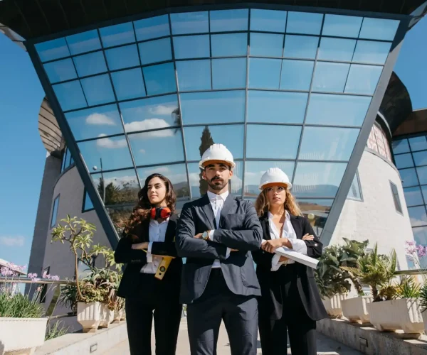Foto de três engenheiros com capacetes de segurança posando na frente de um edifício moderno de vidro, transmitindo confiança e inovação em projetos de construção civil.