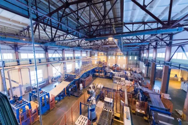 Foto da vista interna de uma planta industrial com estruturas metálicas, tubulações, máquinas e plataformas em um grande galpão, com iluminação e áreas de trabalho ao fundo.