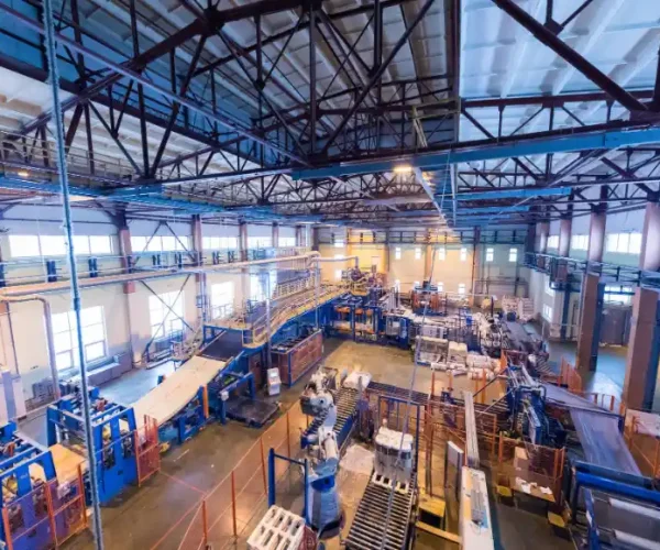 Foto da vista interna de uma planta industrial com estruturas metálicas, tubulações, máquinas e plataformas em um grande galpão, com iluminação e áreas de trabalho ao fundo.