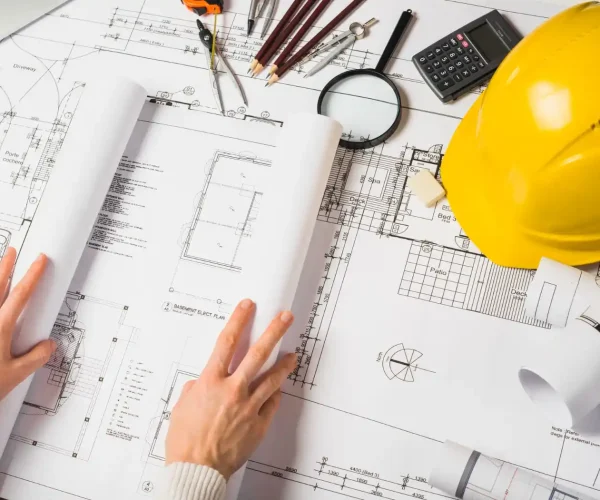 Foto de projetos de construção civil com plantas, ferramentas de desenho, uma calculadora, uma lanterna e um capacete de segurança amarelo.