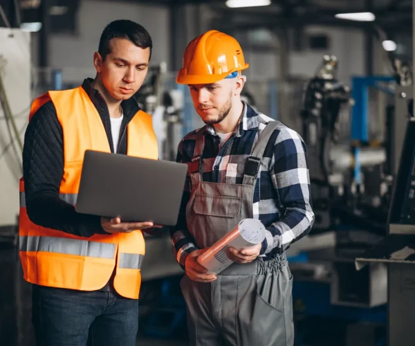 Foto de dois trabalhadores de indústria em ambiente de fábrica usam coletes e capacete de segurança, um deles segura um documento e o outro analisa um notebook.