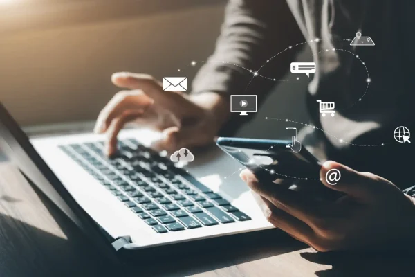 Profissional usando computador portátil e smartphone, com ícones conectados de comunicação digital, representando a abrangência do Inbound Marketing B2B.