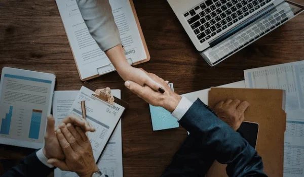 Foto de duas pessoas de gêneros diferentes apertando as mãos em uma mesa de trabalho com laptops, documentos, um contrato assinado e gráficos financeiros ao redor, simbolizando parceria e negócios potencializados com a estratégia de Marketing de Relacionamento B2B.
