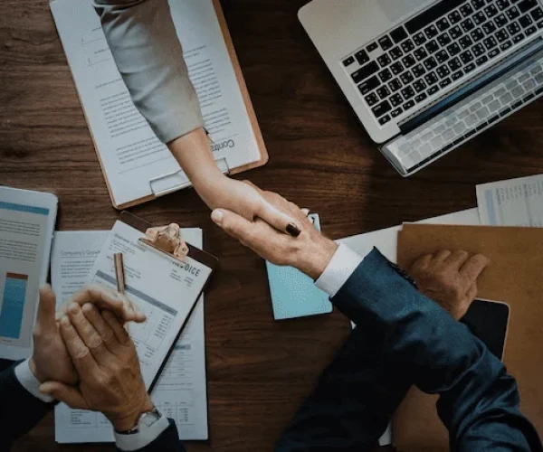 Foto de duas pessoas de gêneros diferentes apertando as mãos em uma mesa de trabalho com laptops, documentos, um contrato assinado e gráficos financeiros ao redor, simbolizando parceria e negócios potencializados com a estratégia de Marketing de Relacionamento B2B.