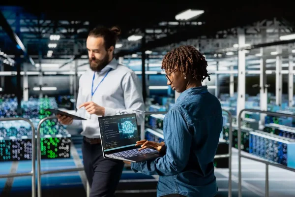 Foto de uma profissional de TI negra analisando um painel com código e outro colaborador conferindo informações em uma prancheta, ambos em um data center.