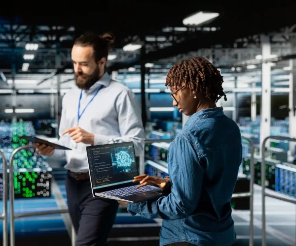 Foto de uma profissional de TI negra analisando um painel com código e outro colaborador conferindo informações em uma prancheta, ambos em um data center.
